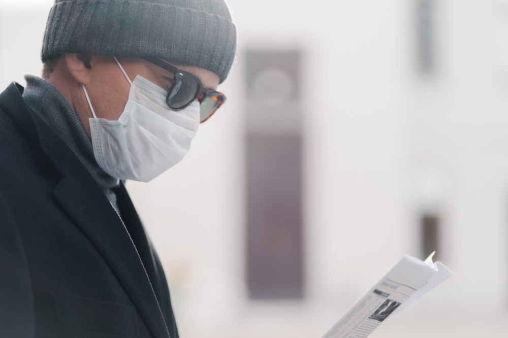 Sideways shot of man in hat, sunglasses and protective mask, reads newspaper outdoor, poses at street, finds out news from press during coronavirus outbreak and quarantine, respiratory virus