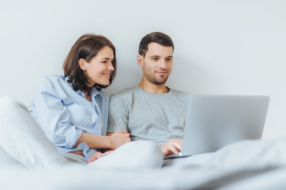 Affectionate female and male have joy together, watch film in bedroom on laptop computer, enjoy togetherness and free internet connection. Lovely couple in love have good time in bedroom