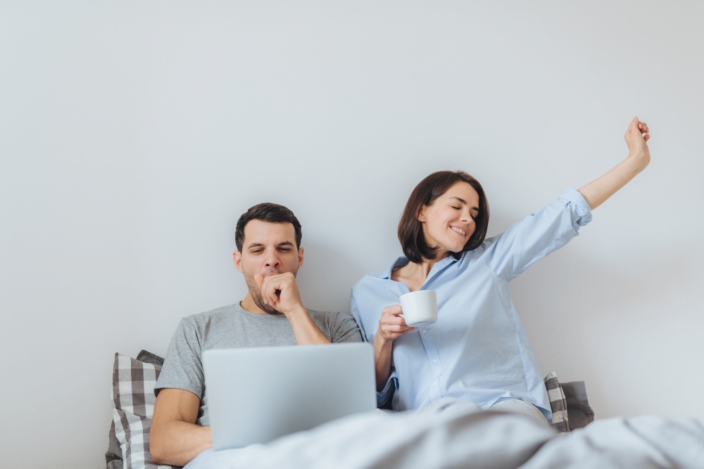 Glad female stretches in bed, drinks hot coffee and her husband works all night on laptop computer, yawns as wants to sleep, spend time together in bedroom, isolated over white background
