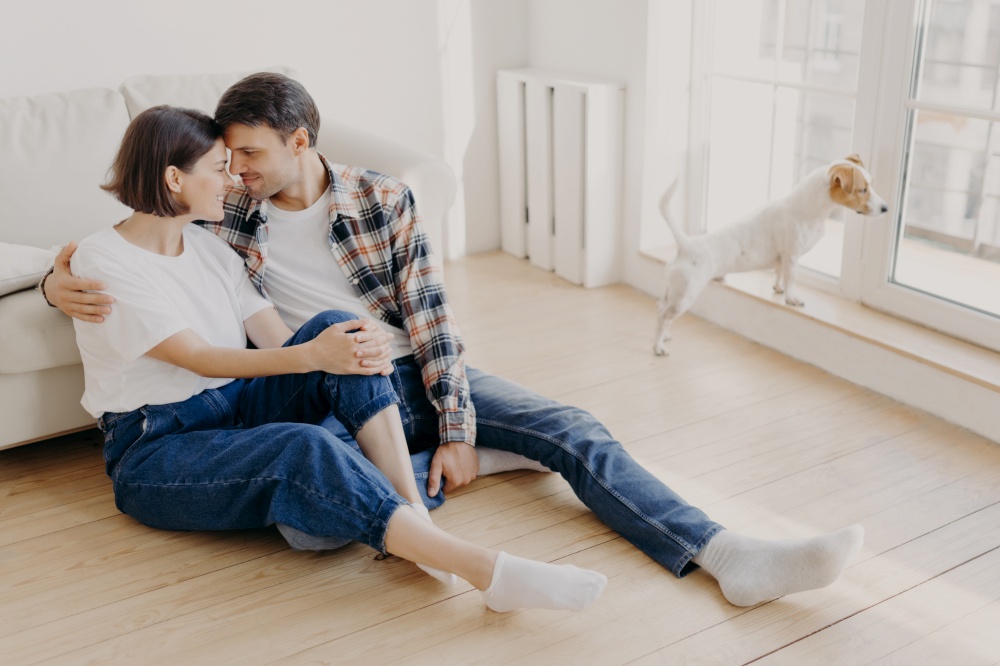 Horizontal shot of pleased husband and wife embrace and kiss, have good relationship, sit on wooden floor near couch, enjoy being at home during weekend, live in new apartment, start new life in abode
