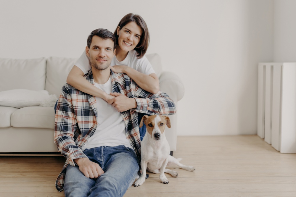 Happy brunette woman embraces husband with love, being in good mood, smiles positively. Husband, wife and dog pose together in living room of new dwelling, enjoy comfort. Couple in love indoor