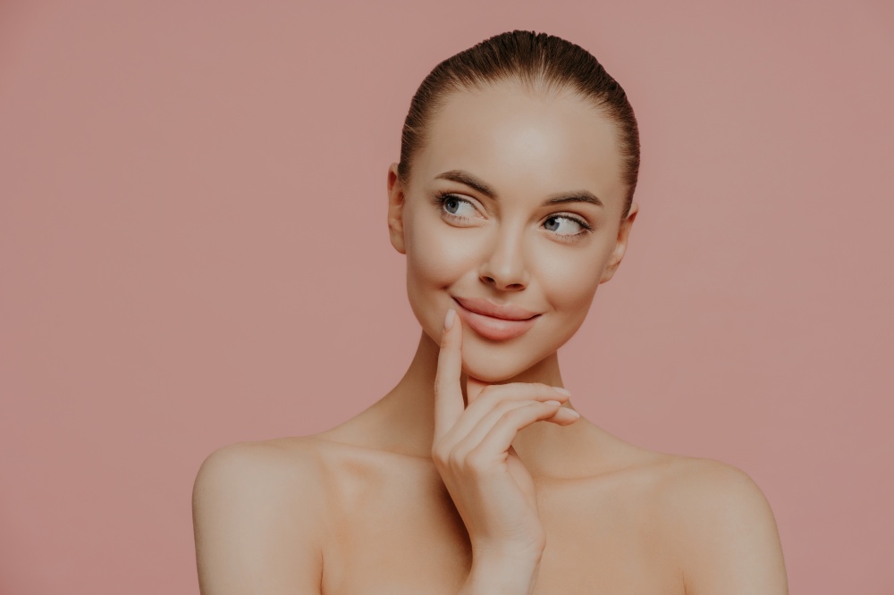 Topless thoughtful lady with clean perfect skin, dark hair, looks aside, undergoes facial trearrments, poses indoor, isolated over rosy background. Cosmetology, skin care and wellness concept