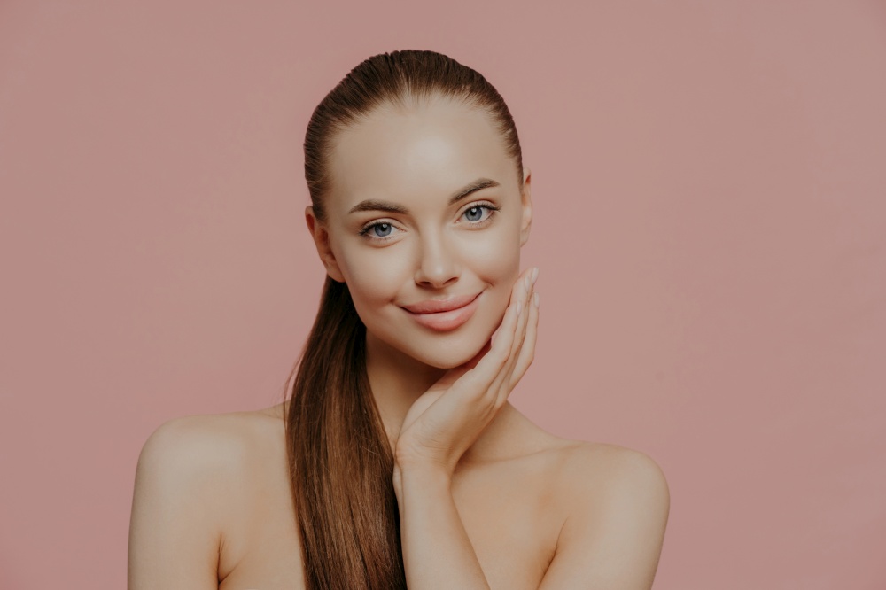 Portrait of good looking female has long straight hair combed in pony tail, stands shirtless, has clean perfect skin, satisfied with her natural beauty, poses indoor against pink background.