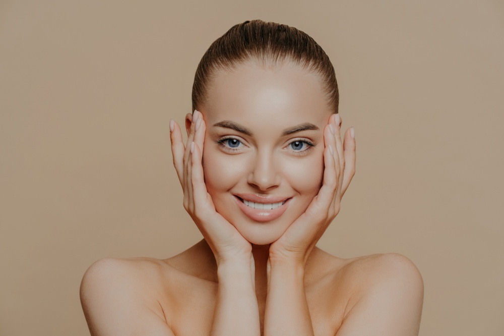 Skin care and beauty concept. Headshot of beautiful cheerful woman touches face gently, has perfect smile, healthy skin after cleaning or applying facial mask, isolated over beige background
