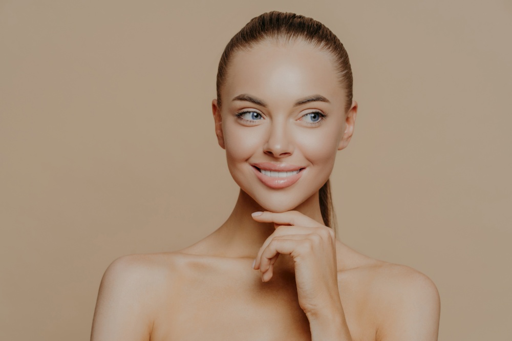 Smiling well groomed young woman with beauty face care, healthy glowing skin, touches chin gently and looks aside, poses shirtless against brown studio background, loves her skin, feels refreshed