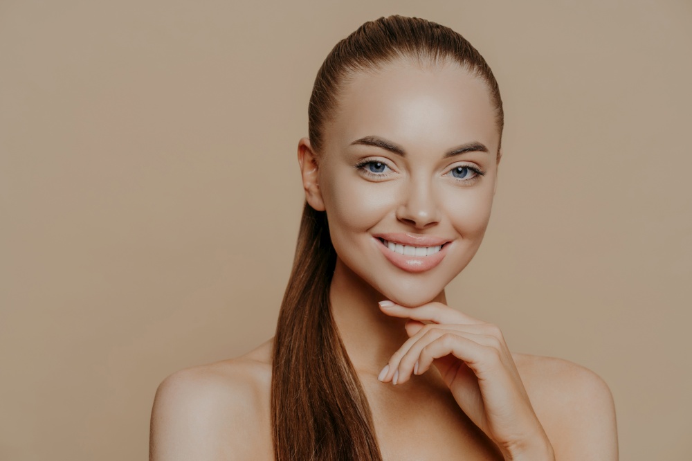 Isolated shot of young woman with European appearance has pure healthy smooth skin after doing daily cleansing procedures, smiles toothily, stands with bare shoulders indoor over beige wall.