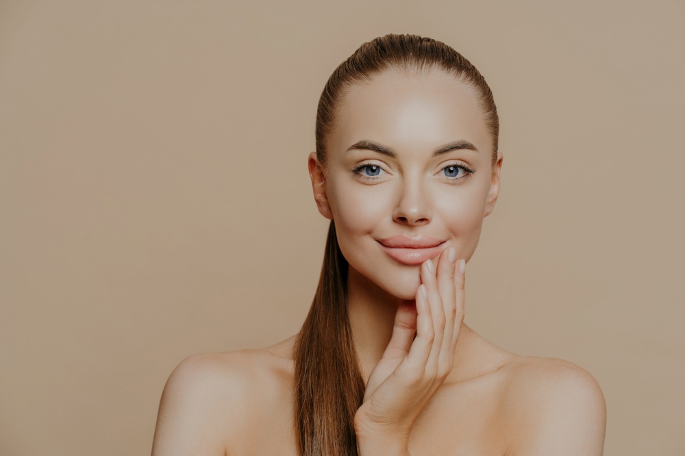 Close up portrait of beautiful naked young woman smiles tenderly and touches face, has healthy clean skin, cares about appearance, poses against beige background. Youth and skin care concept.
