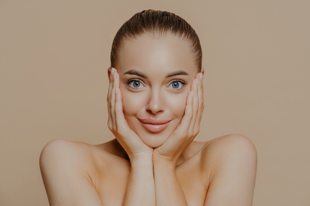 Headshot of young woman with surprised expression, touches face with both hands, has combed hair, natural makeup, healthy soft skin, stands against beige background. Facial treatment, cosmetology, spa