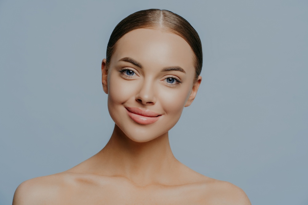 Headshot of attractive woman has beautiful facial features, natural makeup, dark combed hair, naked body, full lips, isolated on blue background, undergoes beauty treatments. Skin care concept