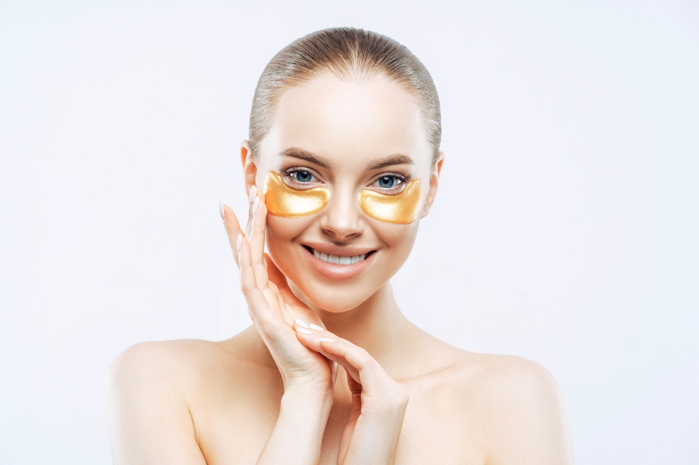 Studio shot of attractive European woman touches face gently, smiles tenderly, applies golden hydrogel patches under eyes, isolated on white background. Eye mask treatment. Anti age procedure