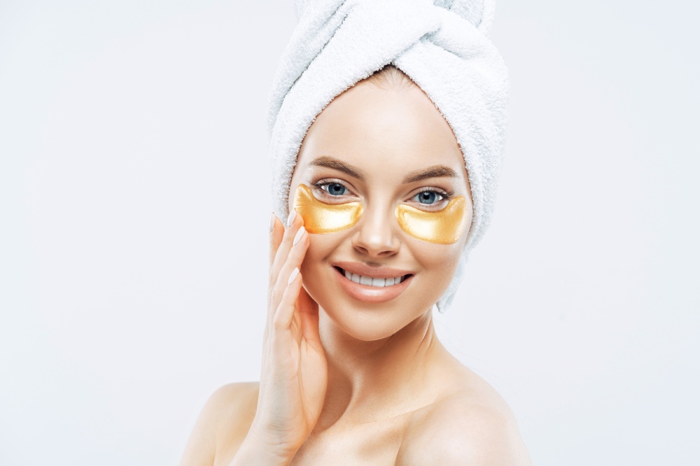Headshot of pretty young European woman has gentle smile, wears golden patches under eyes, cares about face and skin, wears bath towel on head, isolated over white studio wall. Skin care concept