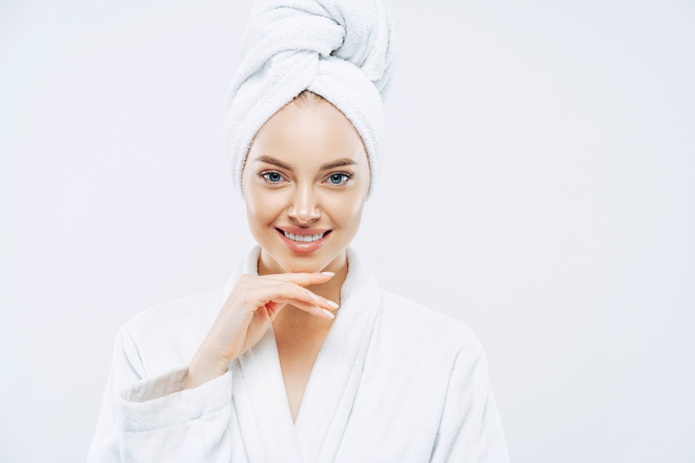 Women, beauty, hygiene, wellness concept. Fresh young European woman wears bath towel and robe, touches chin gently, spends free time in spa, undergoes beauty treatments after shower white background.