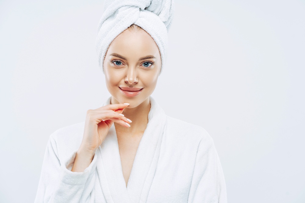 Studio shot of pretty beauty woman has washed hair, wears wrapped towel on head, has manicure, cute natural face, touches chin gently, looks with tender smile, dressed in bath robe, poses indoor