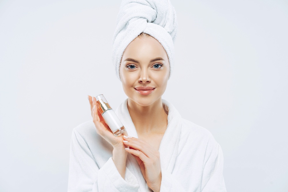 Calm gorgeous woman with healthy skin holds bottle of beauty body lotion for care, poses in bath robe after taking shower, advertises cosmetic prodcut, isolated on white background. Natural beauty