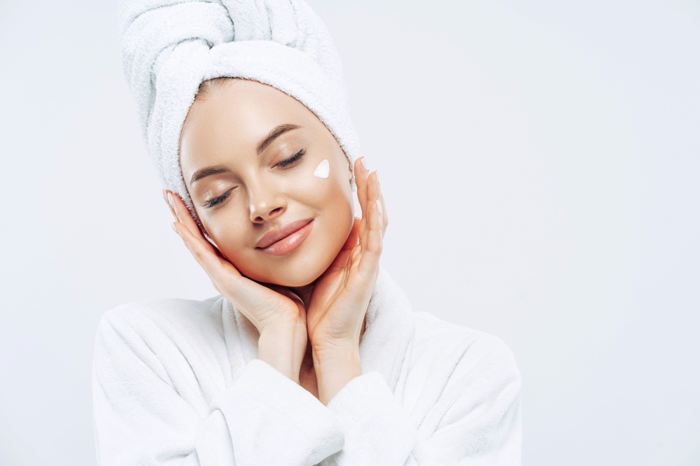 Studio shot of relaxed beautiful woman with closed eyes tilts head, touches skin gently, applies face cream, wears bath towel on head after taking spa procedures, enjoys facial treatments, body care