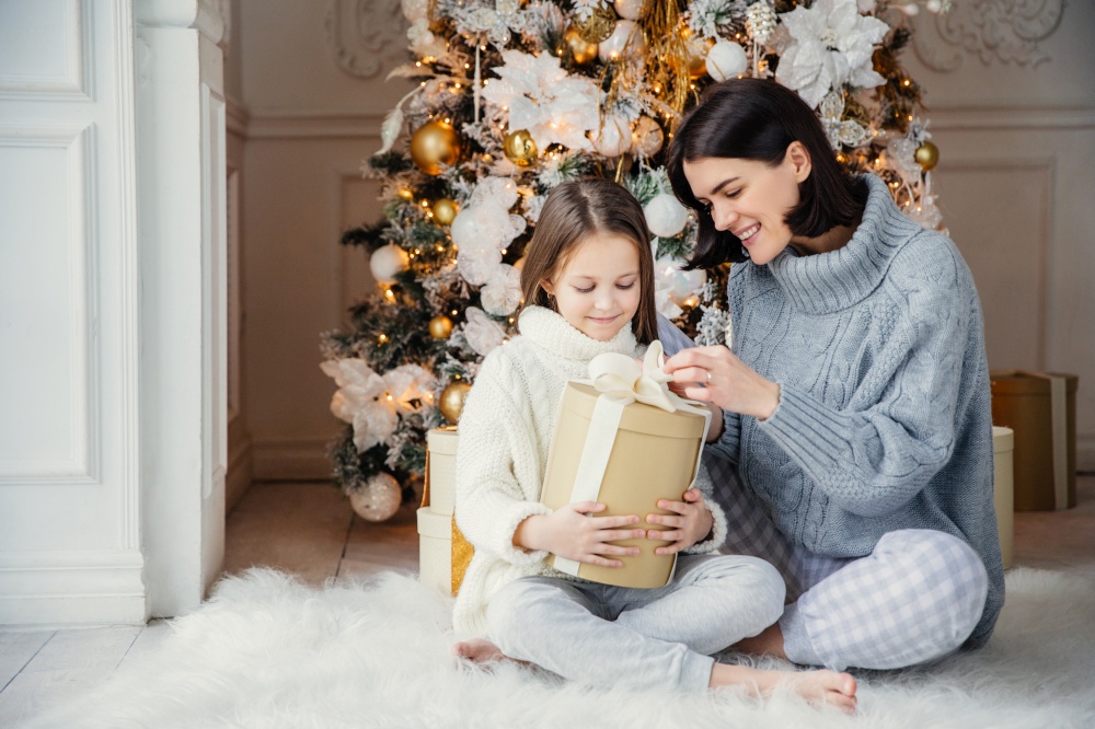 Portrait of pretty girl and beautiful female mother sit together on warm carpet, hold present box, enjoy decorated New Year tree. Family spend time together. Celebration and holidays concept