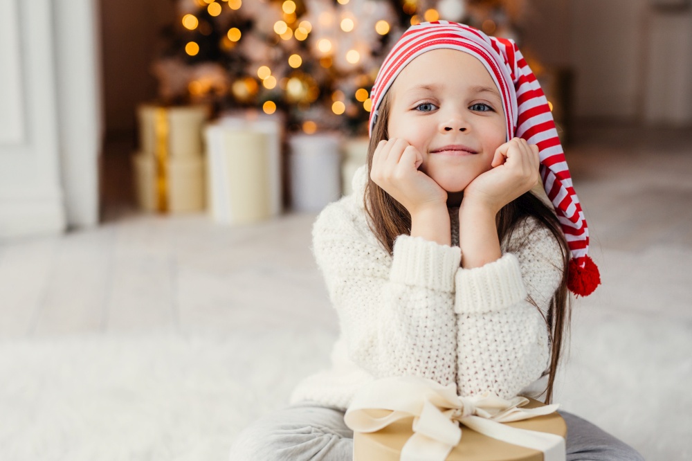 Horizontal portrait of adorable little child, leans on hands with present box, sits against decorated Christmas tree. Blue eyed small kid in knitted white sweater poses at camera. Holidays concept