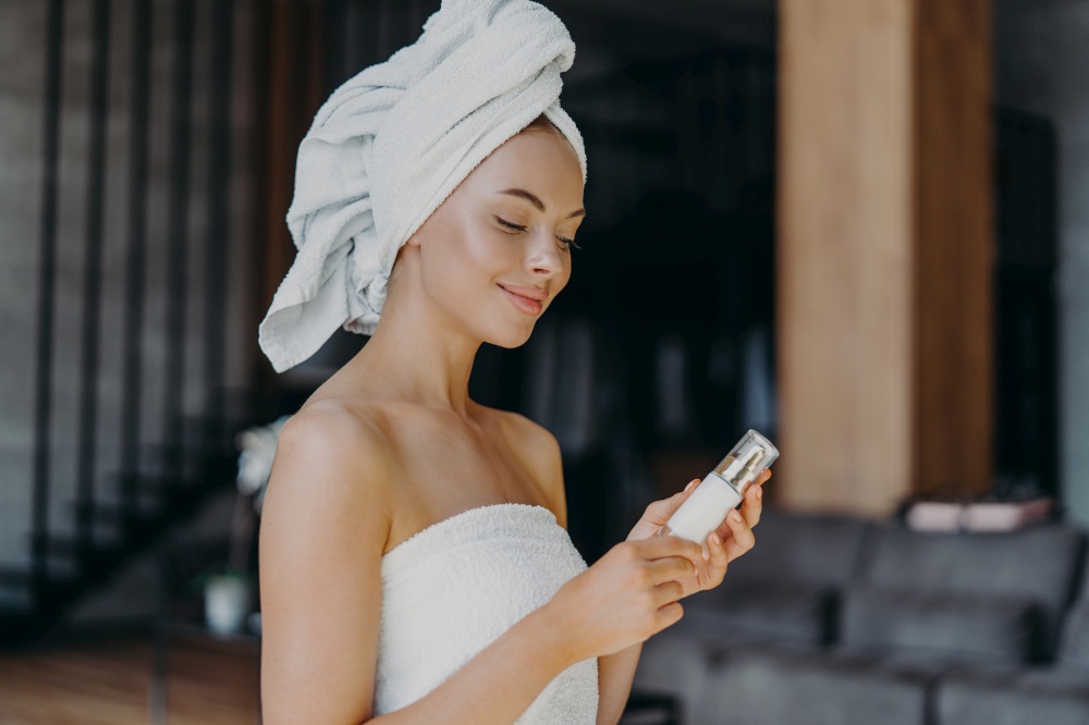 Pleased healthy woman with smooth skin, minimal makeup, holds bottle of body lotion, wrapped in bath towel, poses indoor against blurred background. Female with skin care product. Beauty concept