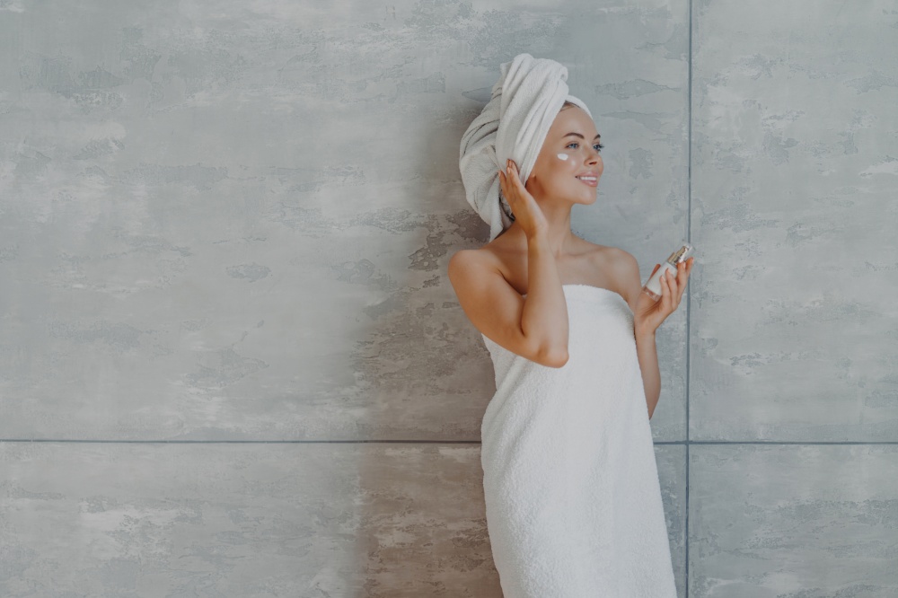 Thoughtful smiling young woman applies foundation or moisturizer on face, holds bottle of body lotion, wrapped in bath towel, concentrated aside with charming smile, isolated on grey background