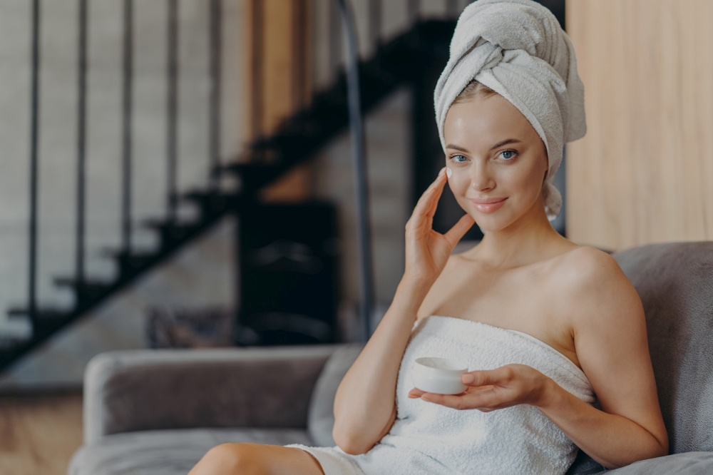 Body care and natural beauty concept. Satisfied blue eyed young woman applies moisturizing cream on face, wears wrapped towel on head, poses on couch against cozy interior, uses good cosmetic