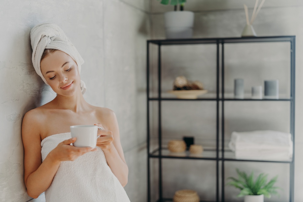 Satisfied healthy European woman poses near wall in bathroom, wrapped in white soft towels, holds mug of tea, being relaxed after spa treatment and taking bath, enjoys hygiene treatments at home