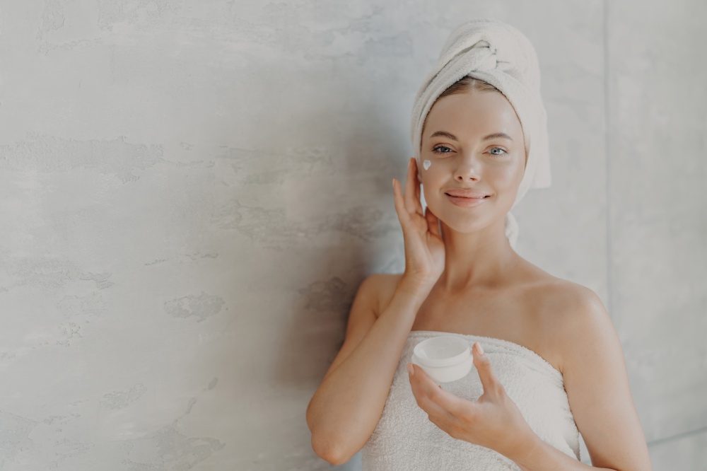 Horizontal shot of lovely female model applies moisturizer on face, holds jar of cosmetic cream has beauty treatment routine, wrapped in bath towel, poses against grey wall with bare shoulders