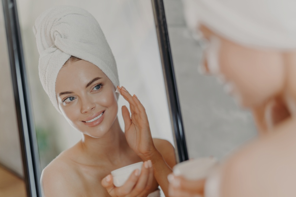 Cute smiling young woman applies beauty cream on face, smiles pleasantly, has minimal makeup, wears wrapped towel on head, looks in mirror, poses in bathroom. Wellness, beauty treatments concept