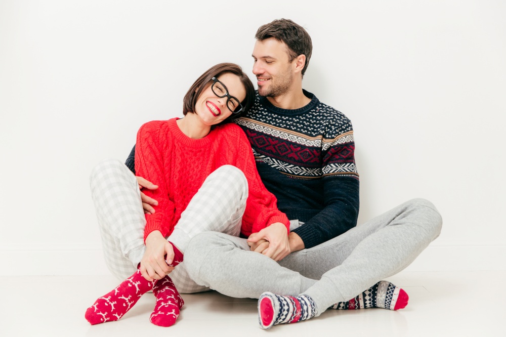 Happy young girlfriend and boyfriend wears sweater and warm socks, sit close to each other on white floor, have positive smiles, being in good mood, have date. Romantic couple cuddle indoor.