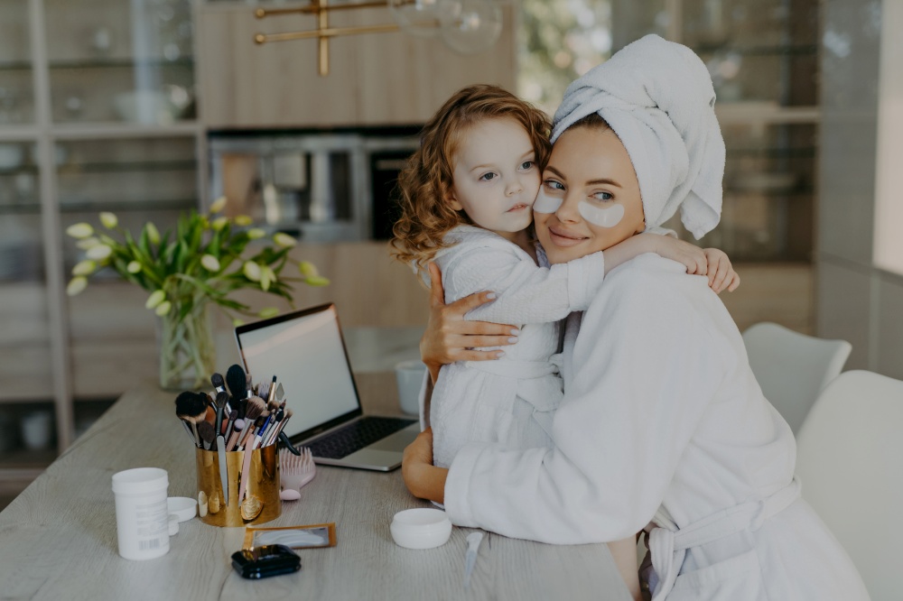 Attractive woman wears hydrogel patches under eyes dressed in bathrobe and wrapped towel embraces her small daughter with love pose together at home near cosmetic tools. Makeup routine concept
