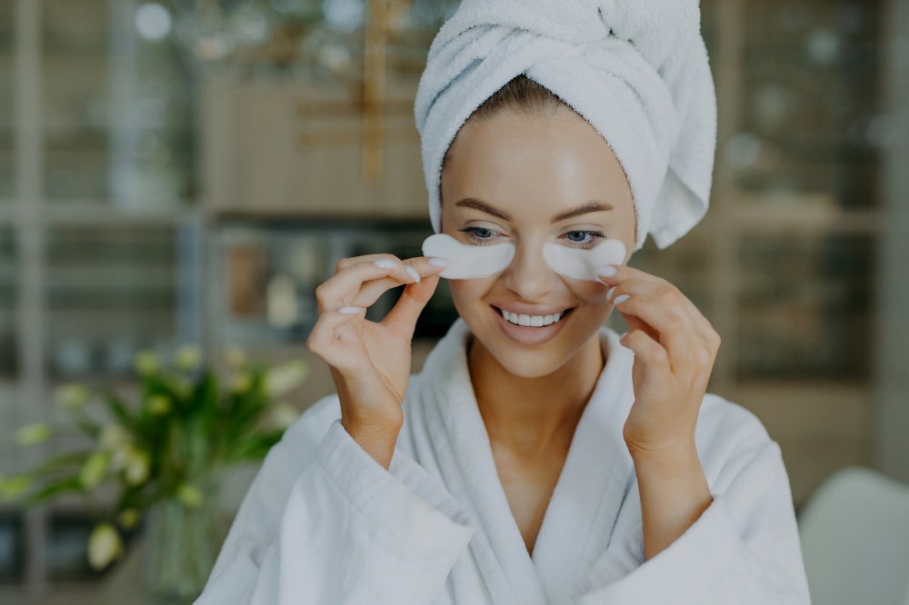 Cheerful young woman with healthy skin takes off beauty patches from under eyes reduces wrinkles or puffiness wears wrapped towel on head and dressing gown smiles toothily has minimal makeup