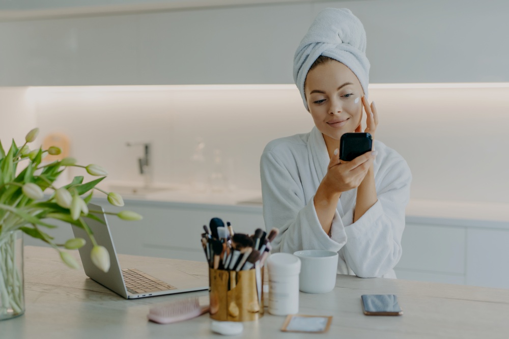 Photo of good looking woman applying face cream on face looks at herself in mirror cares about complexion wears bath towel and robe sits at table against kitchen interior. Beauty time concept