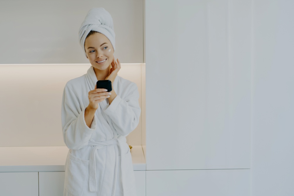 Attractive female applies moisturising cream on face takes care of her complexion and skin holds mirror wears white bathrobe smiles genlty looks thoughtfully poses over modern furniture at home