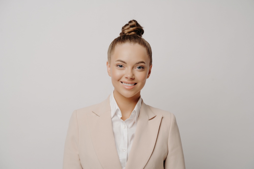 Picture of young attractive business woman happy after conference, female director with hair bun dressed in beige suit and white shirt smiling at camera while standing isolated on grey background. Happy attractive business woman in beige suit