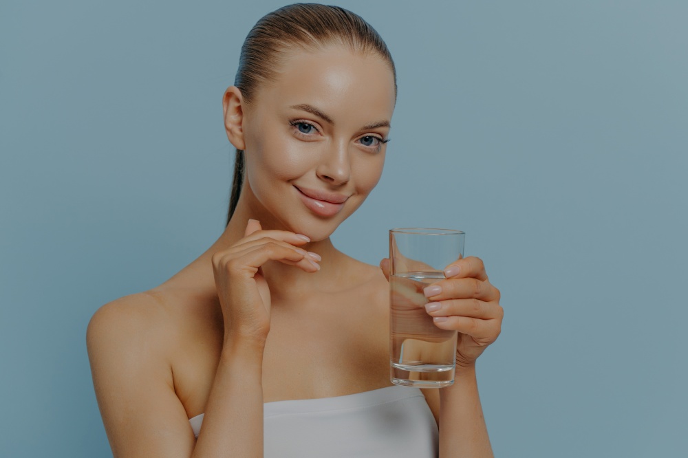 Healthy and hydrated skin. Young charming woman holding glass of pure clean water and touching moisturized facial skin, smiling at camera, posing on blue background. Skincare and healthcare concept. Healthy and hydrated skin. Young charming woman holding glass of clean water and touching face skin