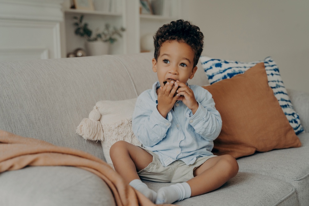 Hungry cute african american boy toddler eating delicious chocolate cupcake that mother brought from local bakery, enjoying the sweet taste while sitting on light sofa in living room at home. Hungry cute afro american boy eating cupcake while spending time at home