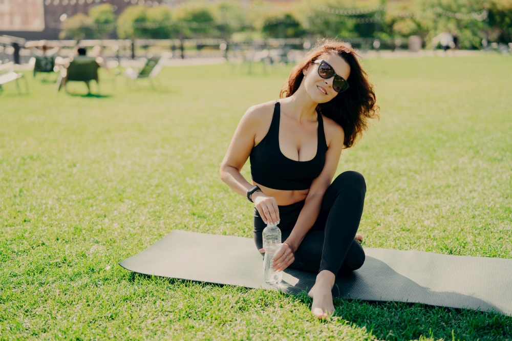 Outdoor shot of brunette slim young woman being in good physical shape trains on fitness mat drinks refreshing water wears sunglasses during sunny day cropped top and leggings. Workout concept