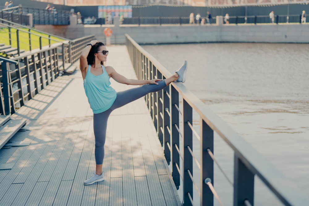 Young fit woman with dark hair stretches legs on fence warms up before jogging wears sunglasses t shirt leggings and sneakers prepares for cardio training poses outdoor being in good physical shape