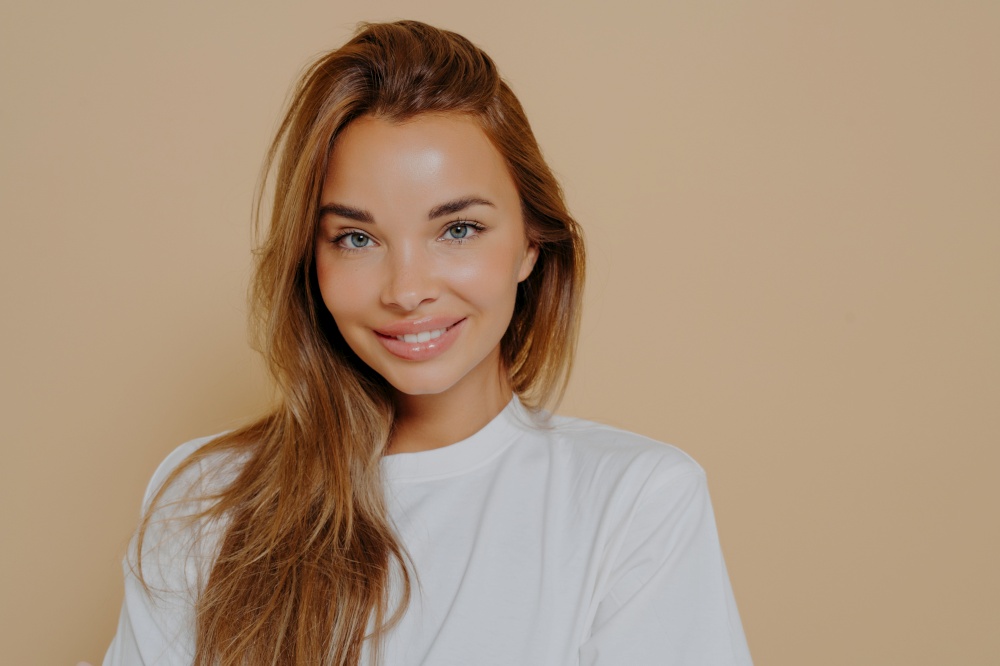 Portrait of beautiful female with long light brown straight hair gently smiling on camera wearing casual white tshirt while poses against pastel beige wall. Women beauty and positive emotions concept. Portrait of smiling beautiful female with long light brown straight hair