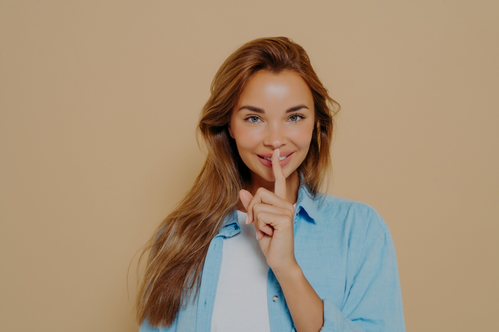 Portrait of joyful smiling female with long light brown hair holding index finger at her lips, saying &rsquo;shh&rsquo;, asking for silent or to keep her secret. Human face expressions, emotions and feelings. Portrait of joyful smiling female with long light brown hair