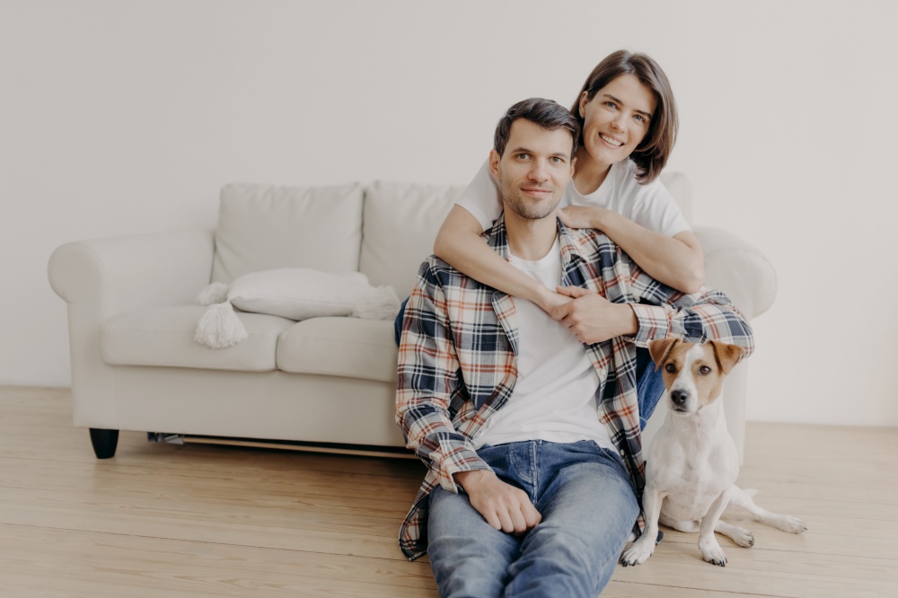 Photo of happy woman embraces her husband, poses on sofa, little dog sits on floor near owner, look all in camera with happy expressions, have romantic relationship. Family portrait. People and home