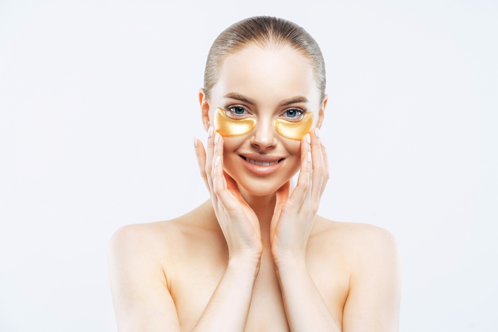 Personal hygiene, beauty, wellness concept. Tender smiling European woman touches face, applies under eye patches, collagen mask for fresh facial skin, stands topless against white background