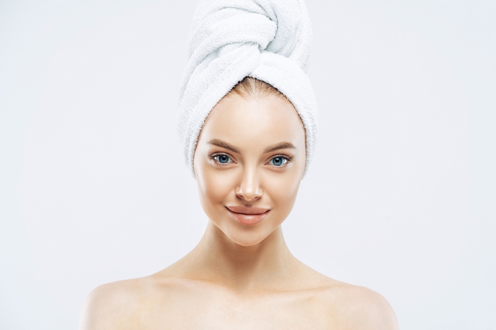 Horizontal shot of charming tender Euoropean woman with healthy skin after taking bath, enjoys hygienic procedures, has natural makeup, wears soft towel on head, stands bare shoulders indoor
