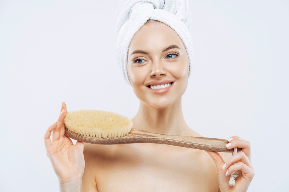 Young woman with healthy fresh skin, uses body brush, smiles gently, wears bath towel on head, poses topless, isolated over white background. Spa accessories. Women, body care, hygiene concept