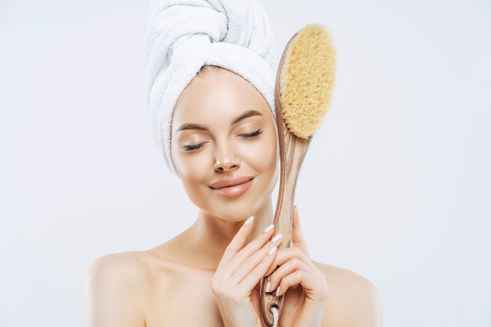 Horizontal shot of pleased female stands with eyes closed, holds big bath brush for massaging skin, enjoys hygienic treatments at home, isolated on white background. Beauty, care, exfoliation