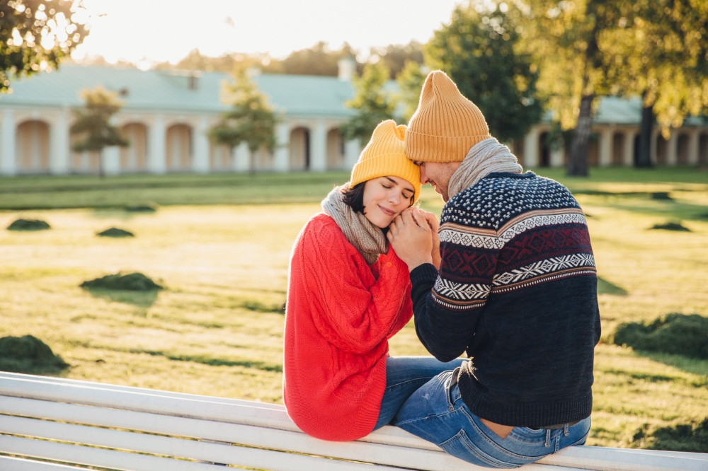 True feelings and romantisism concept. Adorable young woman in knitted yellow hat and red warm sweater warms her hands in boyfriend`s hands, sit together on bench, closes eyes with great enjoyment