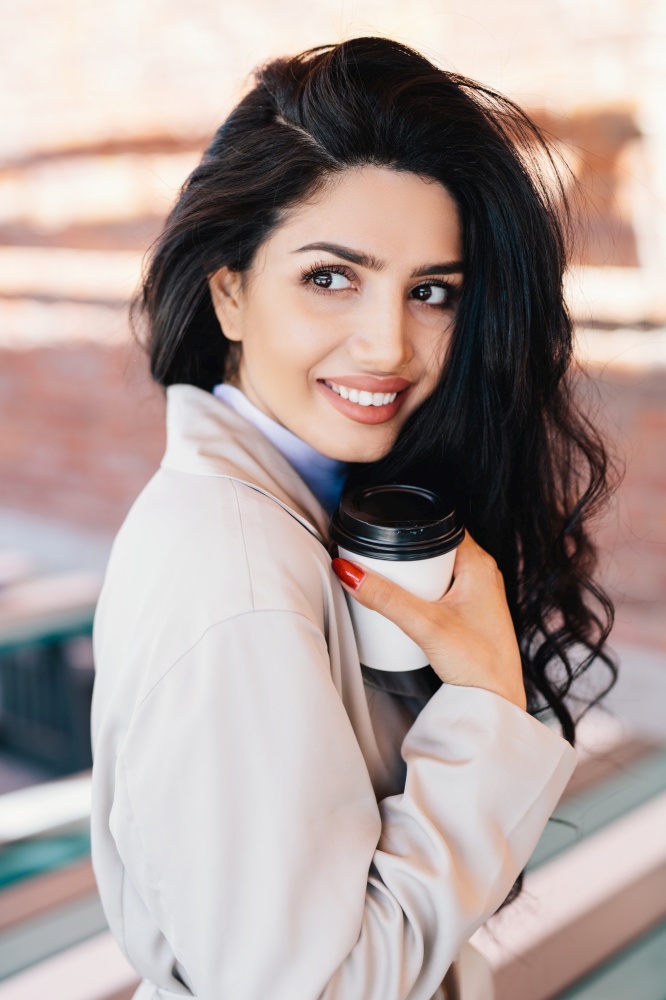 Attractive young woman drinking hot drink from disposable paper cup having perfect luxurious dark hair and red manicure wearing white coat looking aside with delightful expression having rest outdoors