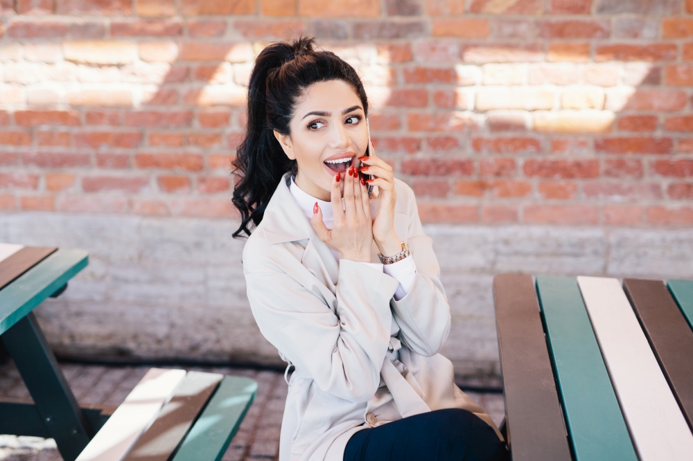 Horizontal portrait of happy female with dark hair and beautiful appearance dressed formally holding her hand on lower lip looking pleased and excited while speaking with her lover over smartphone