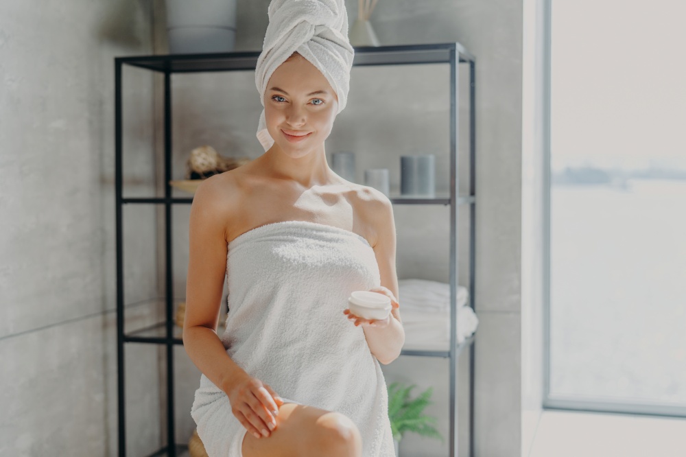 Horizontal shot of pleased smiling woman wrapped in bath towel, touches legs, has silky skin, uses cosmetic product after taking shower, has epilation procedure. Female applies moisturising cream