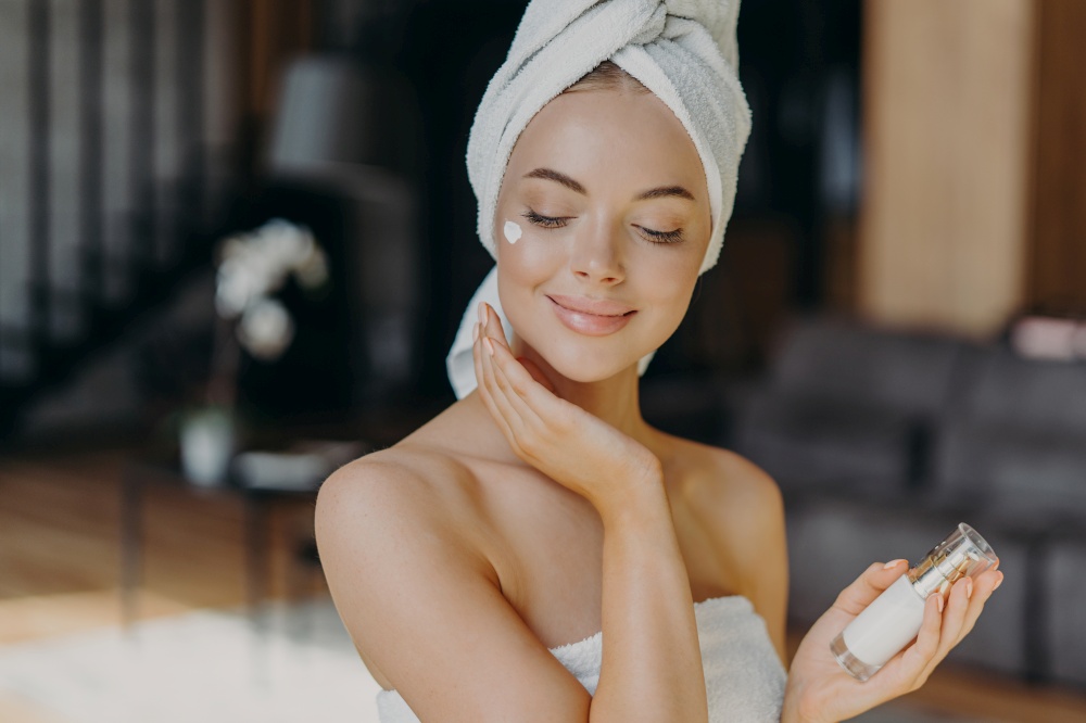 Close up shot of young feminine lady applies face lotion, wears wrapped towel on head, has bare shoulders, smiles with satisfaction, uses cosmetic product. Natural beauty and cosmetology concept