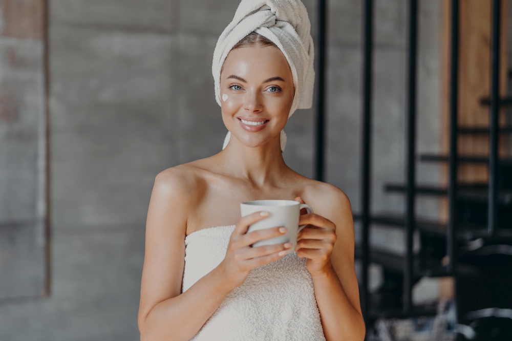 Attractive smiling young woman with makeup, toothy smile, poses bare shoulders, wrapped in towel, drinks hot beverage stands indoor has cheerful expression applies moisturizing cream on face